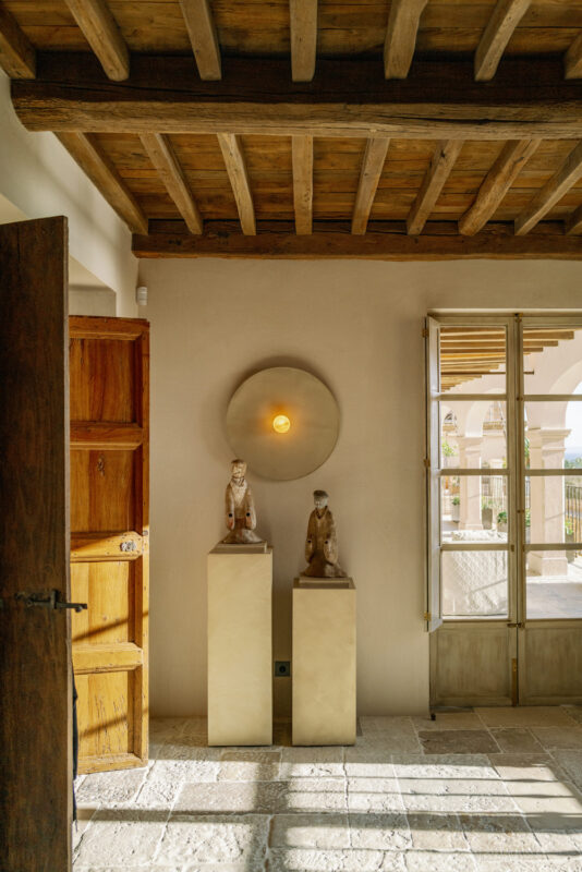 Bright living room corner featuring two sculptures on pedestals, wooden door, and a round light fixture on the wall.
