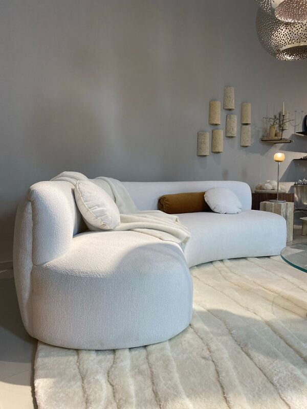 Curved white sofa with cushions on a light rug, set against a neutral wall with decorative elements.