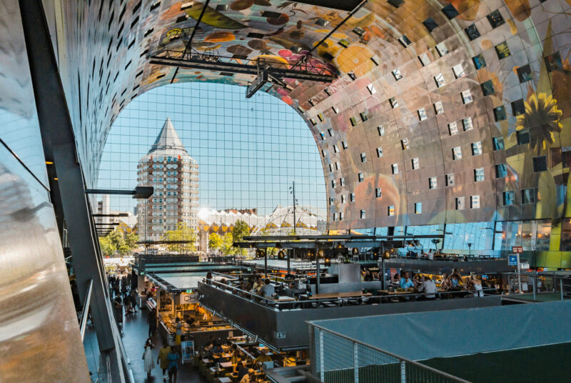 Interior view of a modern market hall with colorful ceiling, reflecting buildings outside, and bustling food stalls below.