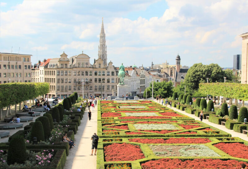 View of a landscaped garden with colorful flower beds, people strolling, and historic buildings in the background.