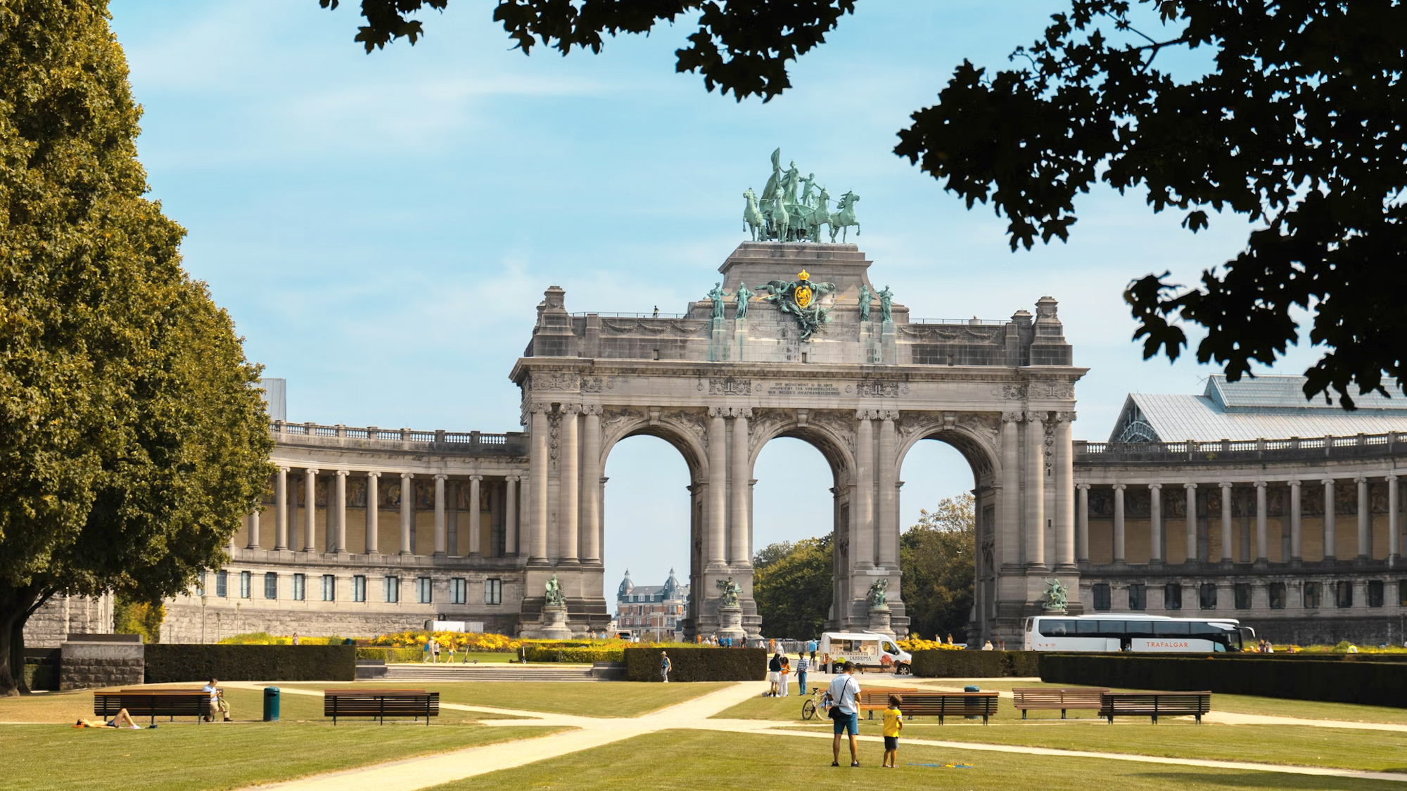 Triumphal arch in a park with people walking, surrounded by greenery and buildings under a clear blue sky.