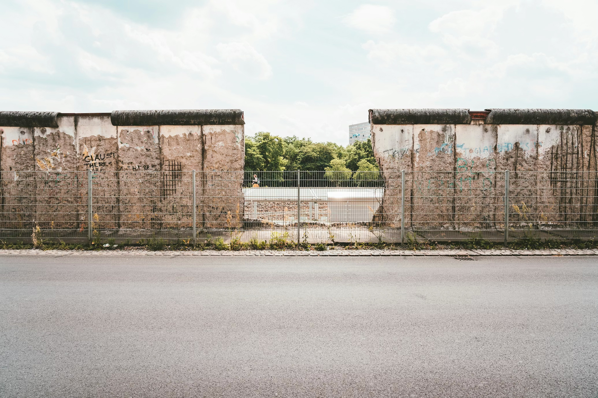 Fragmented concrete wall with graffiti, surrounded by a metal fence, under a cloudy sky.