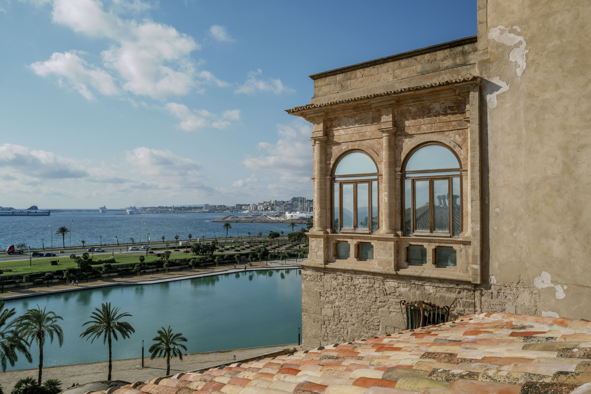 View from a rooftop terrace overlooking a waterfront with palm trees and distant cityscape under a partly cloudy sky.