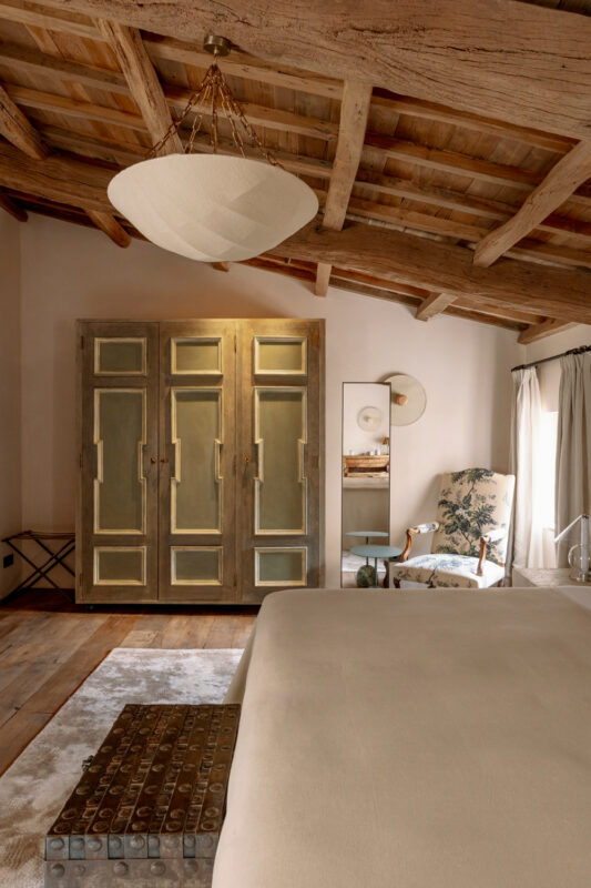 Cozy bedroom featuring a wooden ceiling, a large wardrobe, an armchair, and a bedside table with decorative items.
