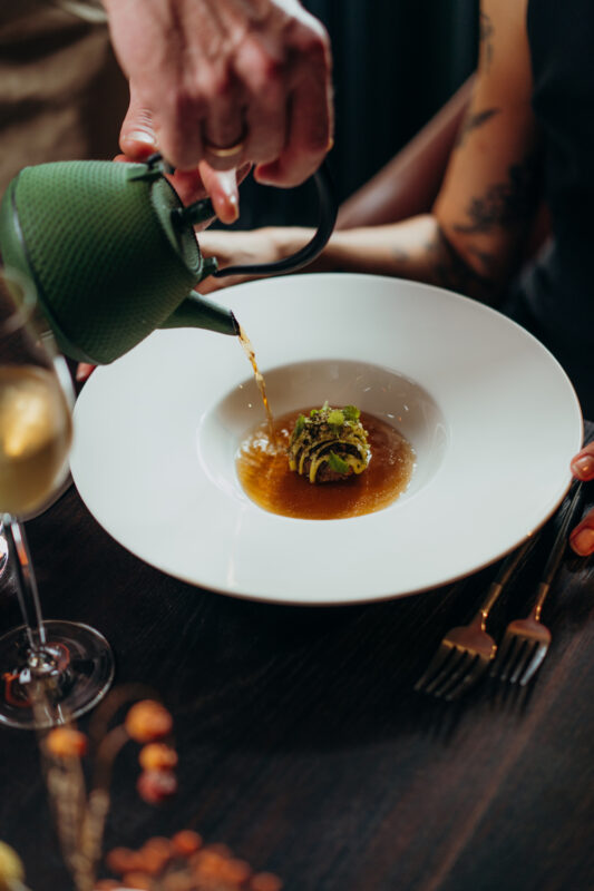 A hand pours broth from a green teapot into a white bowl containing a small dish, with a glass of wine nearby.