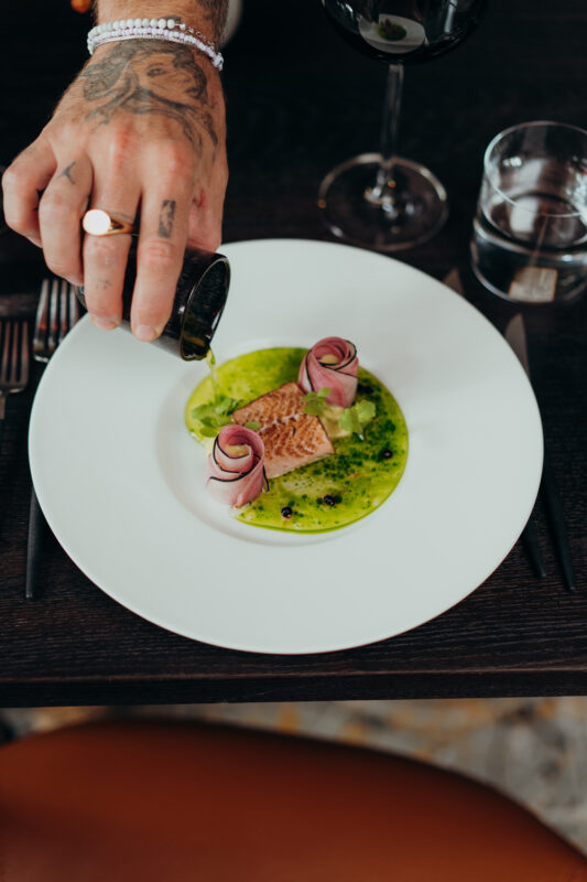 Elegant dish featuring seared meat garnished with rose-shaped vegetables, drizzled with sauce, on a white plate.
