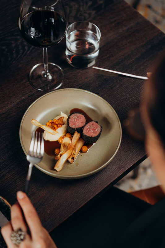 Plate of sliced meat with sauce, accompanied by parsnip fries and a garnish, with a glass of red wine and water nearby.