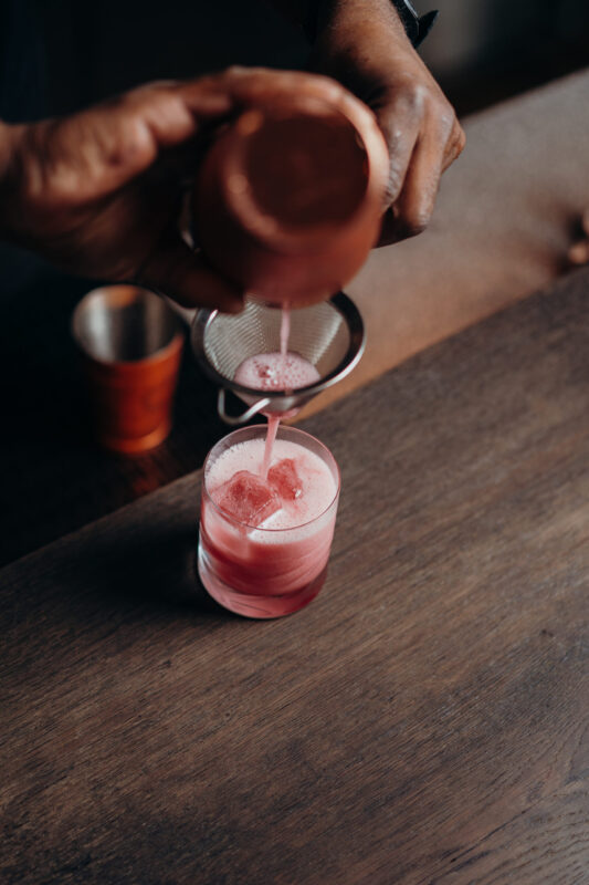 A hand pours a pink cocktail through a strainer into a glass with ice, set on a wooden table.