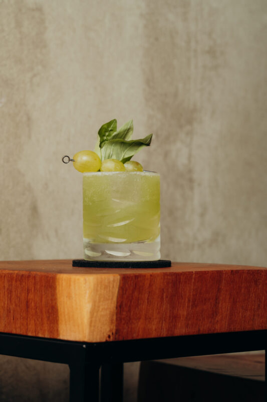 Green cocktail garnished with grapes and basil, served in a glass on a wooden table against a textured wall.