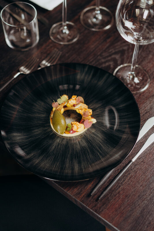 Artfully arranged dish on a black plate, featuring colorful garnishes and a green element, set on a wooden table.