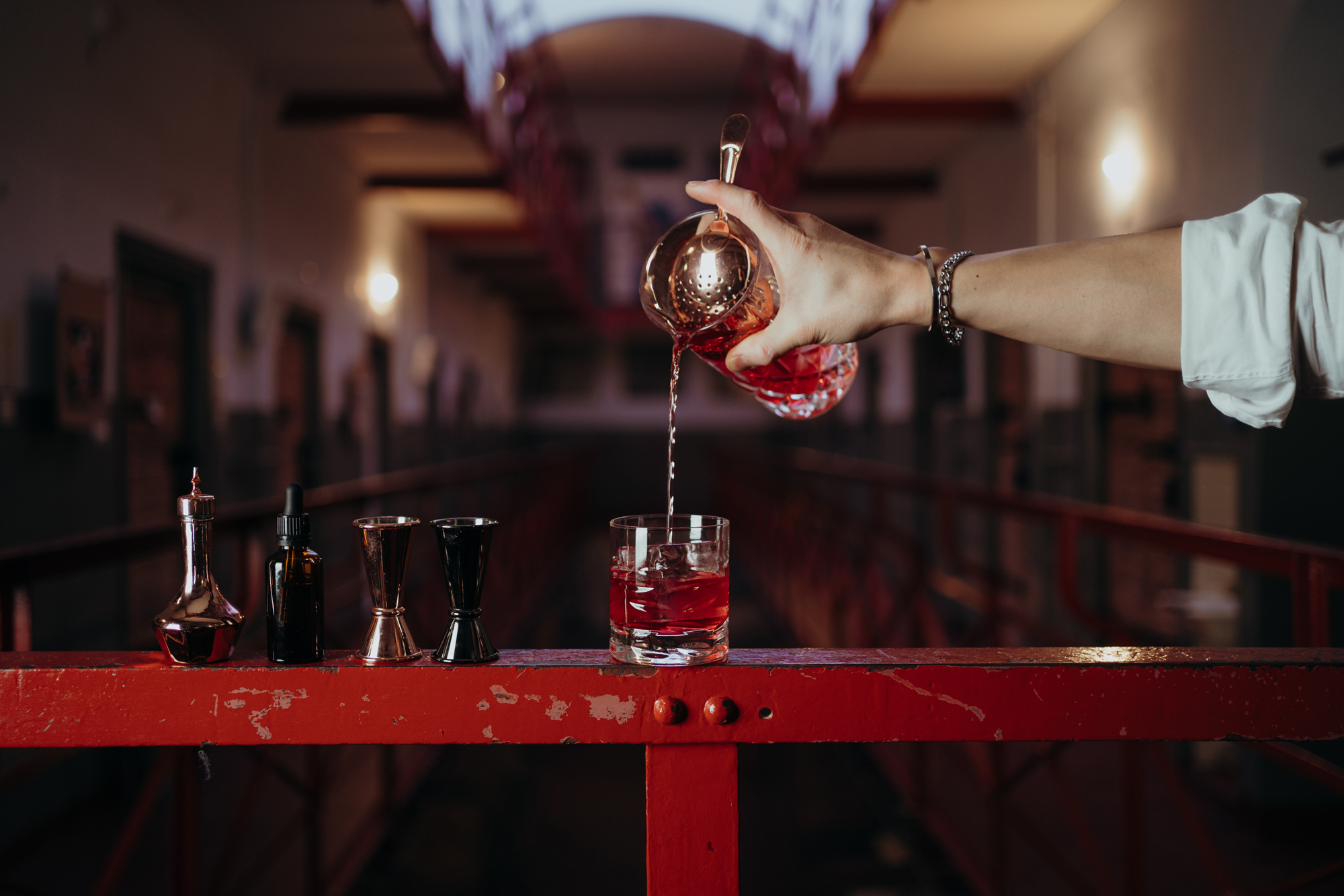 A hand pours a vibrant red cocktail into a glass, with various bar tools on a red railing in a dimly lit corridor.
