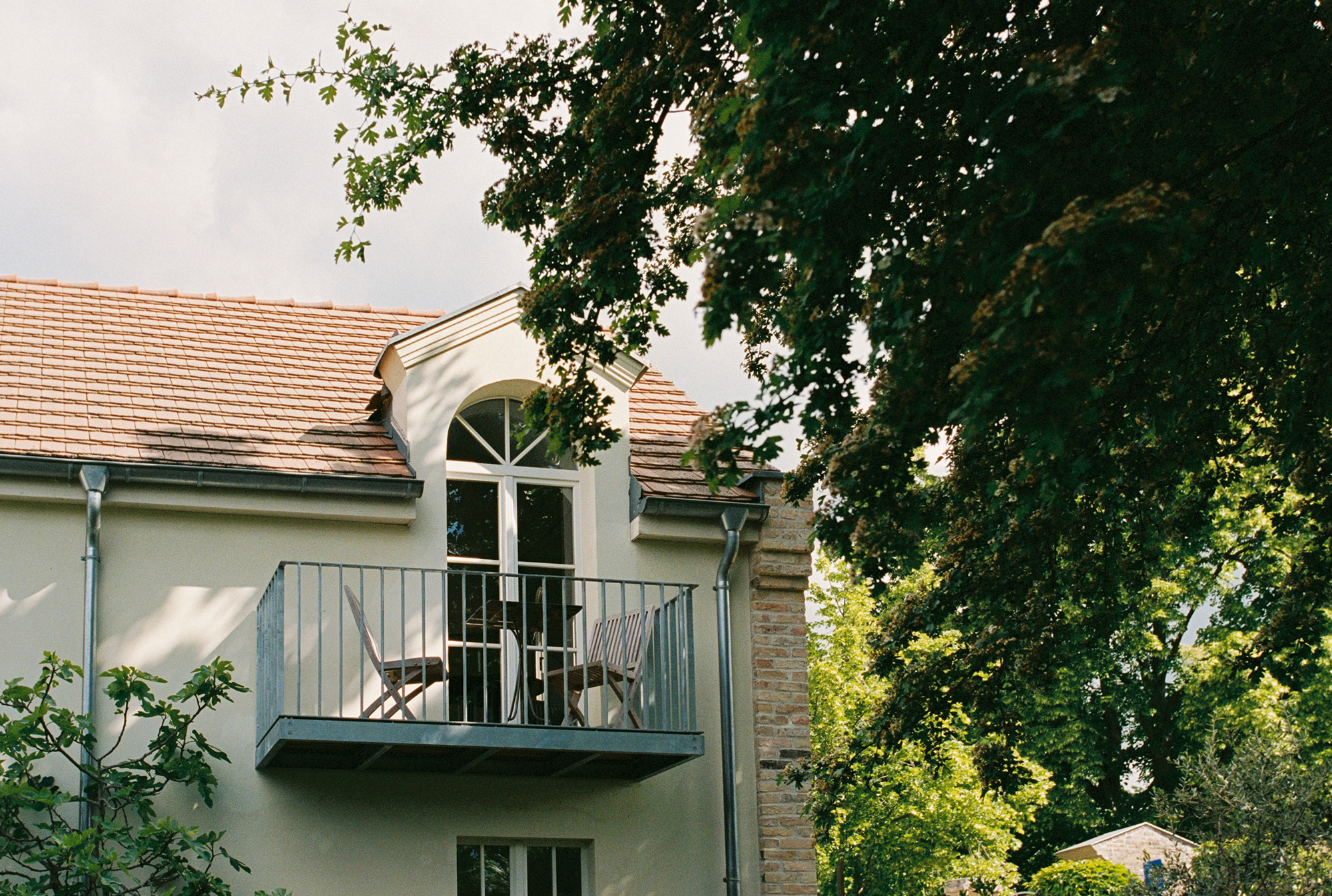 Balcony of a light-colored building with two chairs, surrounded by trees and a cloudy sky.