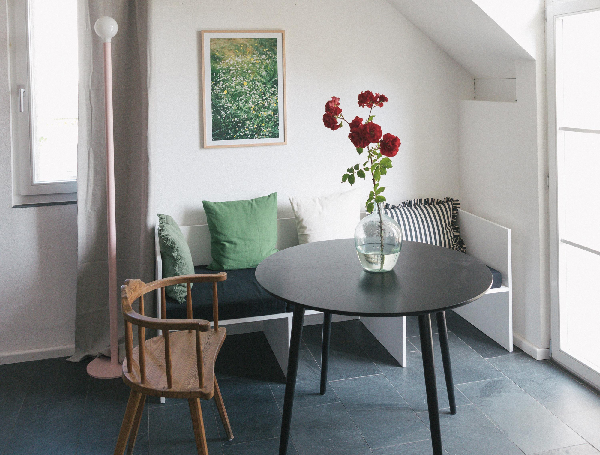 Bright interior featuring a round black table, a wooden chair, and a vase of red roses on the table, with green cushions o...