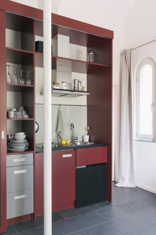 Modern kitchen with red cabinetry, gray drawers, and a black fridge, featuring dishes and glassware on shelves.