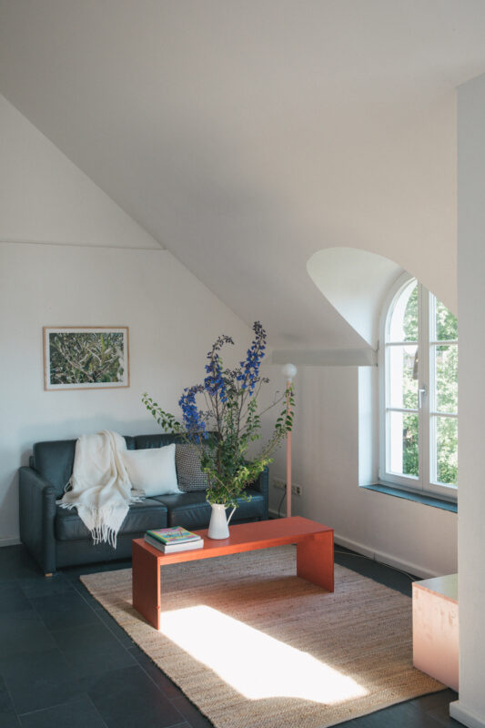 Cozy living area with a dark sofa, orange coffee table, and a vase of blue flowers, featuring large windows and natural li...