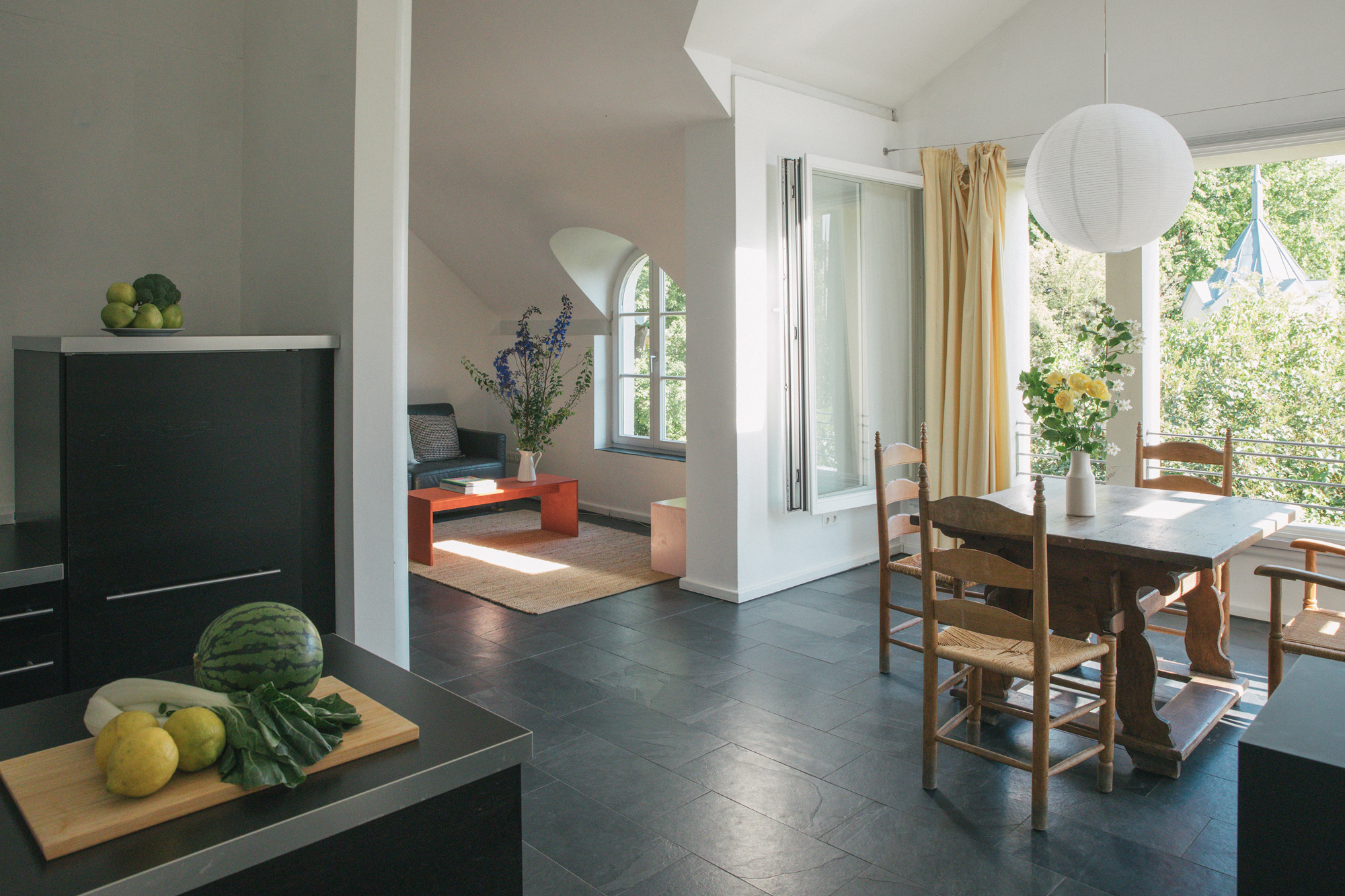 Bright kitchen and dining area with a wooden table, chairs, and large windows overlooking greenery.