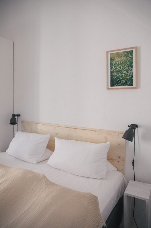 Cozy bedroom with a wooden headboard, two white pillows, a beige blanket, and a framed nature print on the wall.