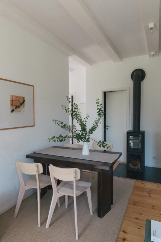 Modern dining area with a wooden table, three light chairs, and a vase of greenery, featuring a black stove in the backgro...