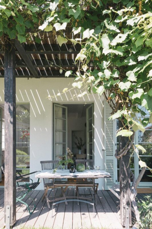 A cozy outdoor dining area with a table set for two, surrounded by green vines and sunlight filtering through.