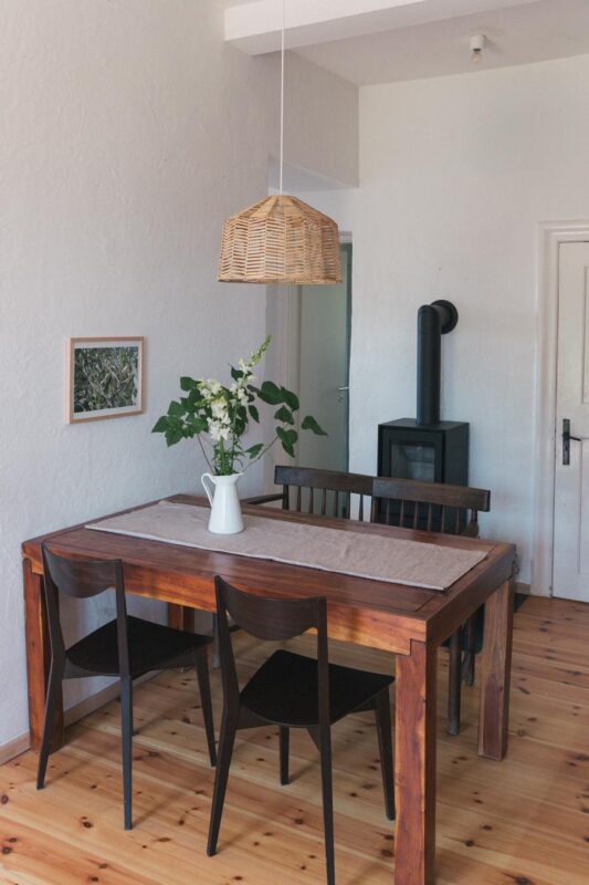 Wooden dining table with black chairs, a vase of flowers, and a wicker pendant light in a bright, minimalist room.