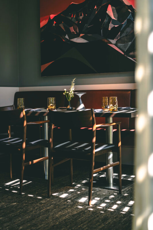 Modern restaurant interior featuring wooden chairs, a table set with glassware, and a large abstract mountain painting.