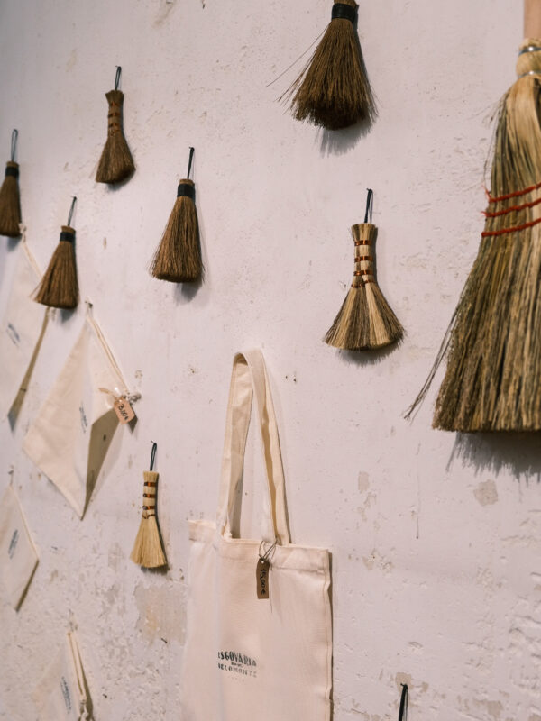 Broom-shaped decorations hang on a textured wall, alongside fabric bags with "Escovaria de Belomonte" branding.