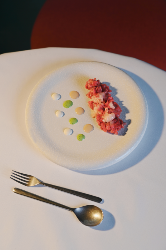 Steak tartare with scampi, garnished with sauces, served on a textured white plate with cutlery beside it.