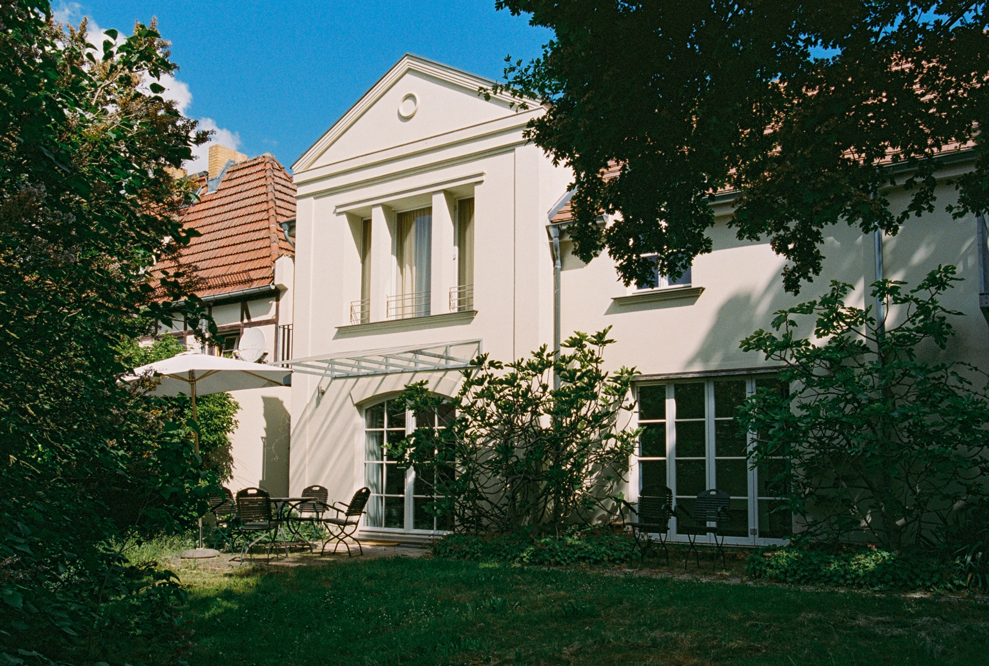 Modern white building with large windows, surrounded by greenery and a patio with chairs and an umbrella.