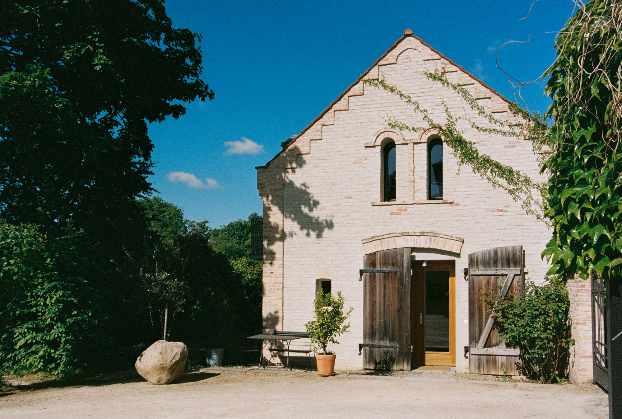Historic brick building with wooden doors, surrounded by greenery and a large rock, under a clear blue sky.