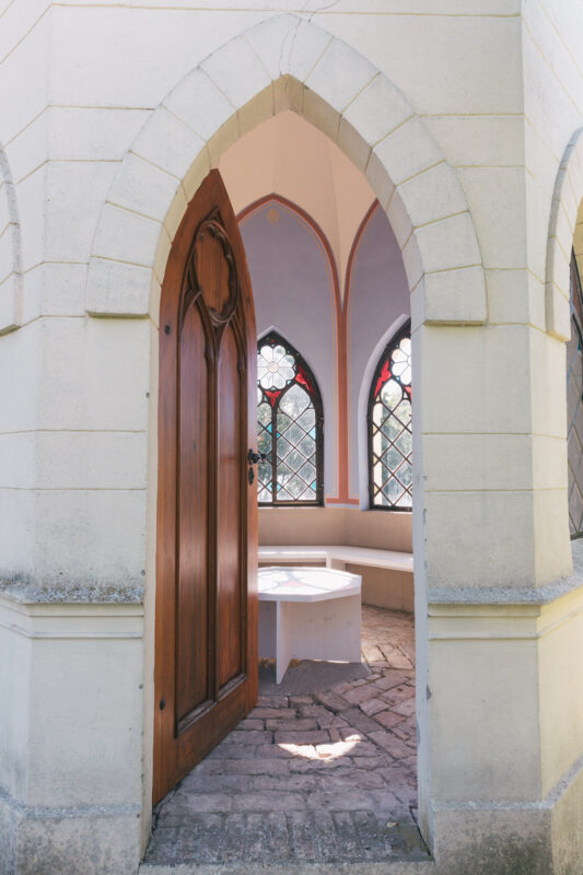 Wooden door with an archway, leading into a room with stained glass windows and a circular bench inside.