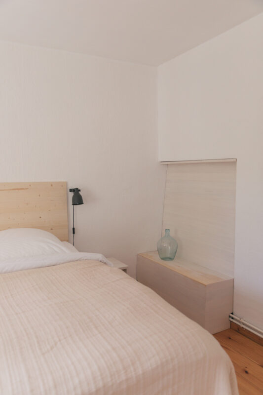Minimalist bedroom featuring a wooden bed with a light-colored blanket, a bedside table, and a glass vase.