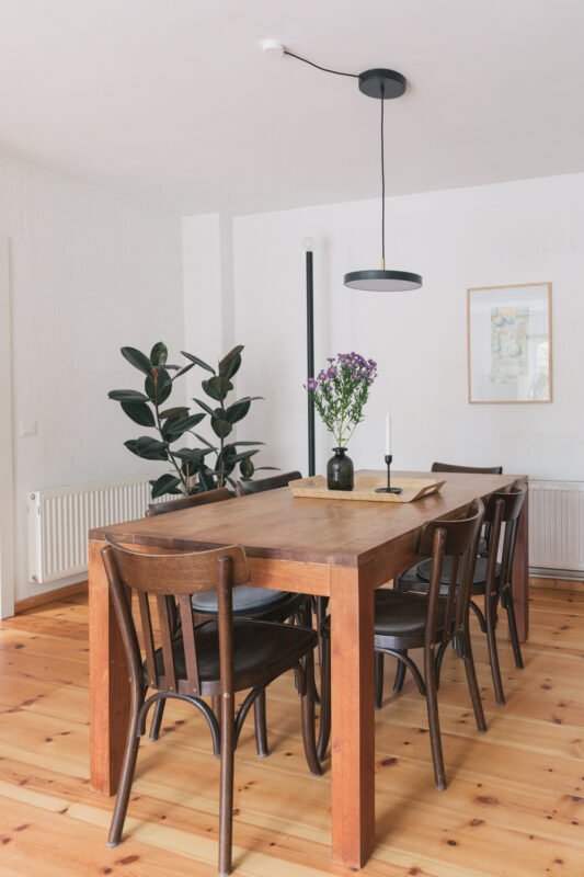 Modern dining room featuring a wooden table with six black chairs, a vase of flowers, and a plant in the corner.