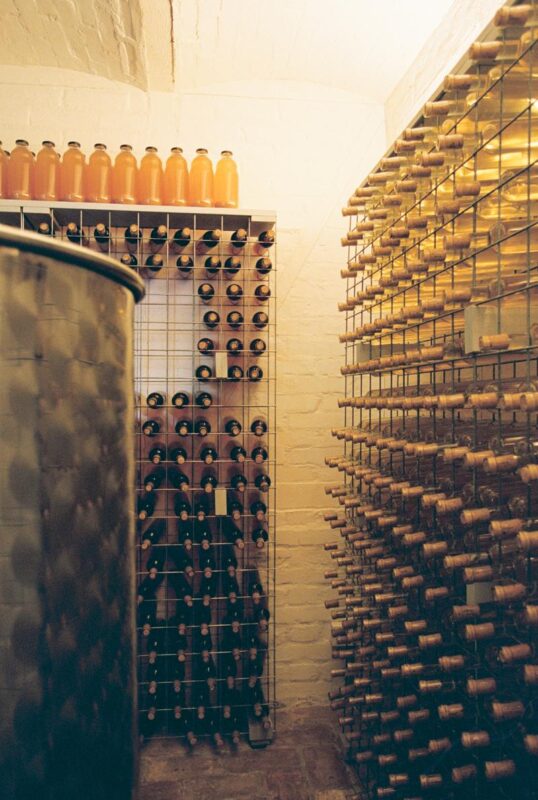 Wine cellar with metal racks holding various bottles, including orange juice bottles on a shelf above.