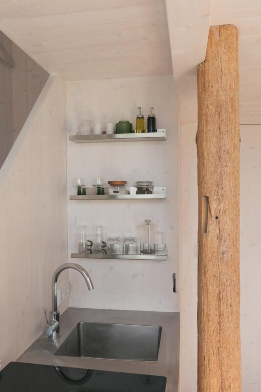 Modern kitchen corner with a stainless steel sink, wooden post, and shelves displaying glassware and condiments.