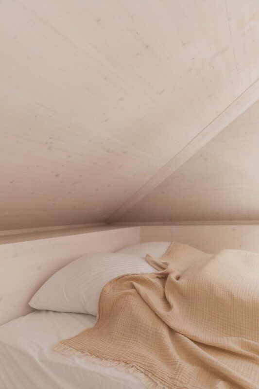 Cozy bedroom corner with a bed featuring white linens and a beige blanket, under a light wooden slanted ceiling.