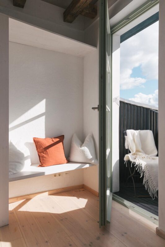 Bright corner of a room with a bench, orange and white pillows, and an open door leading to a balcony with a chair and bla...