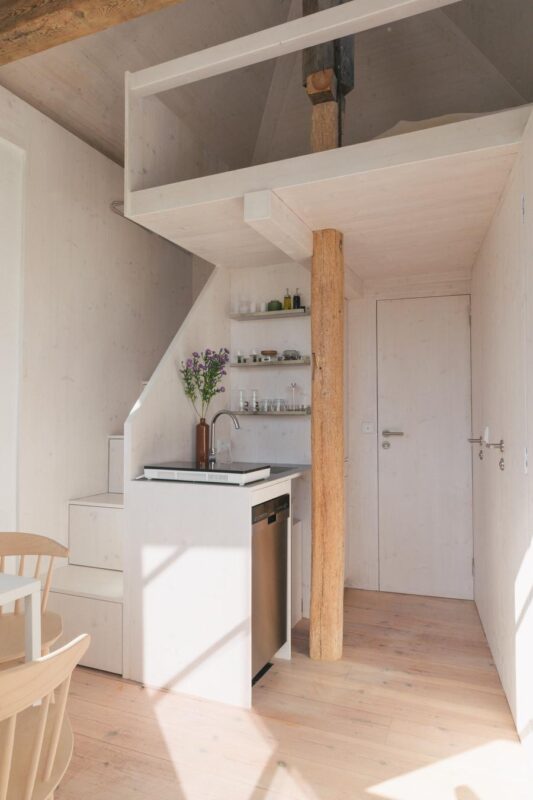 Bright, modern interior of a vacation rental with a kitchenette, wooden beams, and a staircase, featuring a vase of flowers.