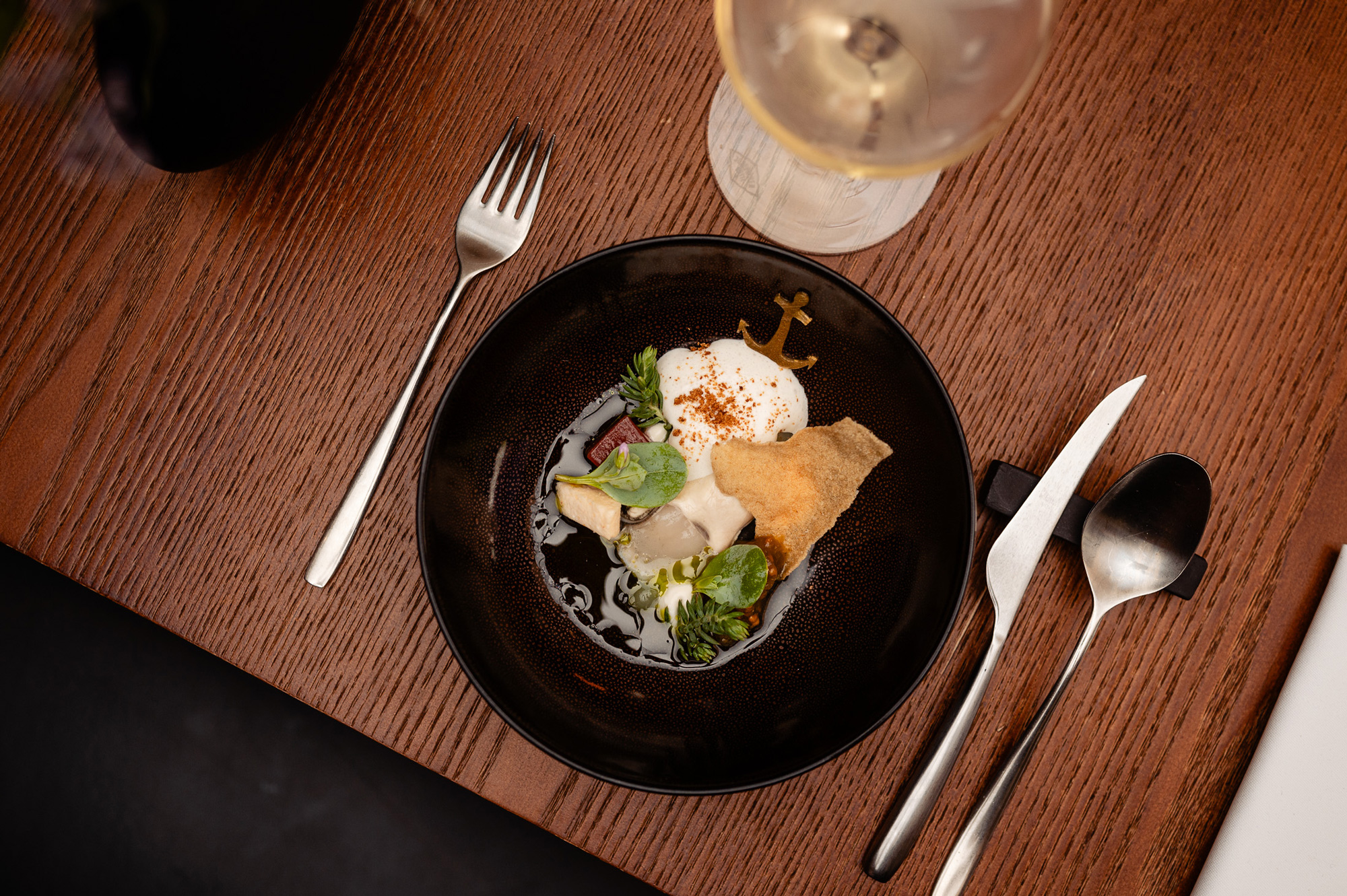 A black plate with a poached egg, oyster, and watermelon, garnished with greens, beside a wine glass and cutlery.