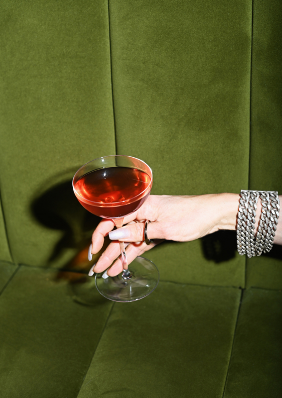 A hand holding a cocktail glass filled with a reddish drink, set against a green velvet background.