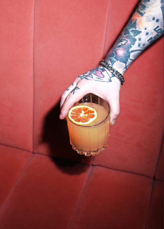 Hand holding a cocktail with an orange slice, resting against a red velvet couch.