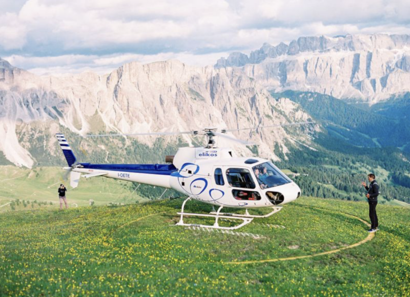 Helicopter on a grassy hill with mountains in the background; two people nearby, one taking a photo.