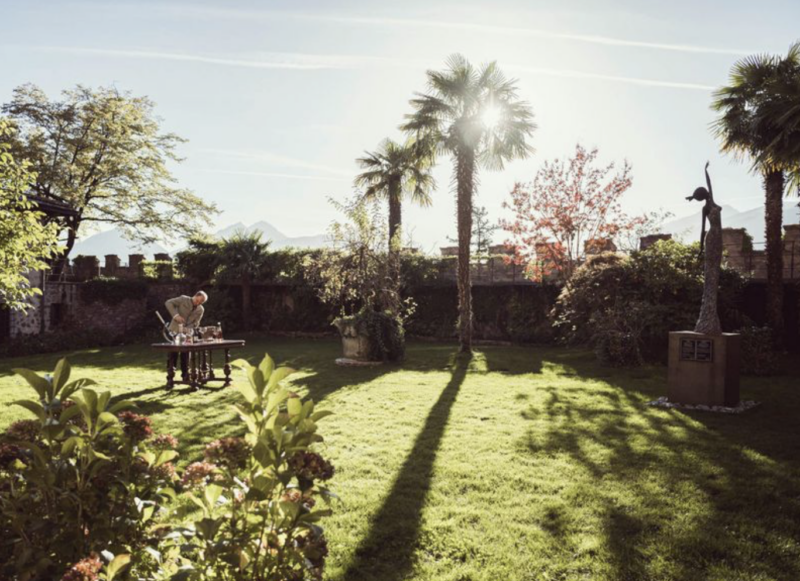 A person sets a table in a sunlit garden surrounded by palm trees and colorful foliage, with mountains in the background.