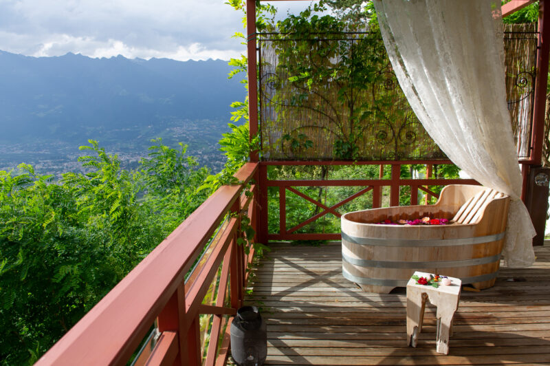 Wooden hot tub on a balcony with a scenic mountain view, surrounded by greenery and a curtain.