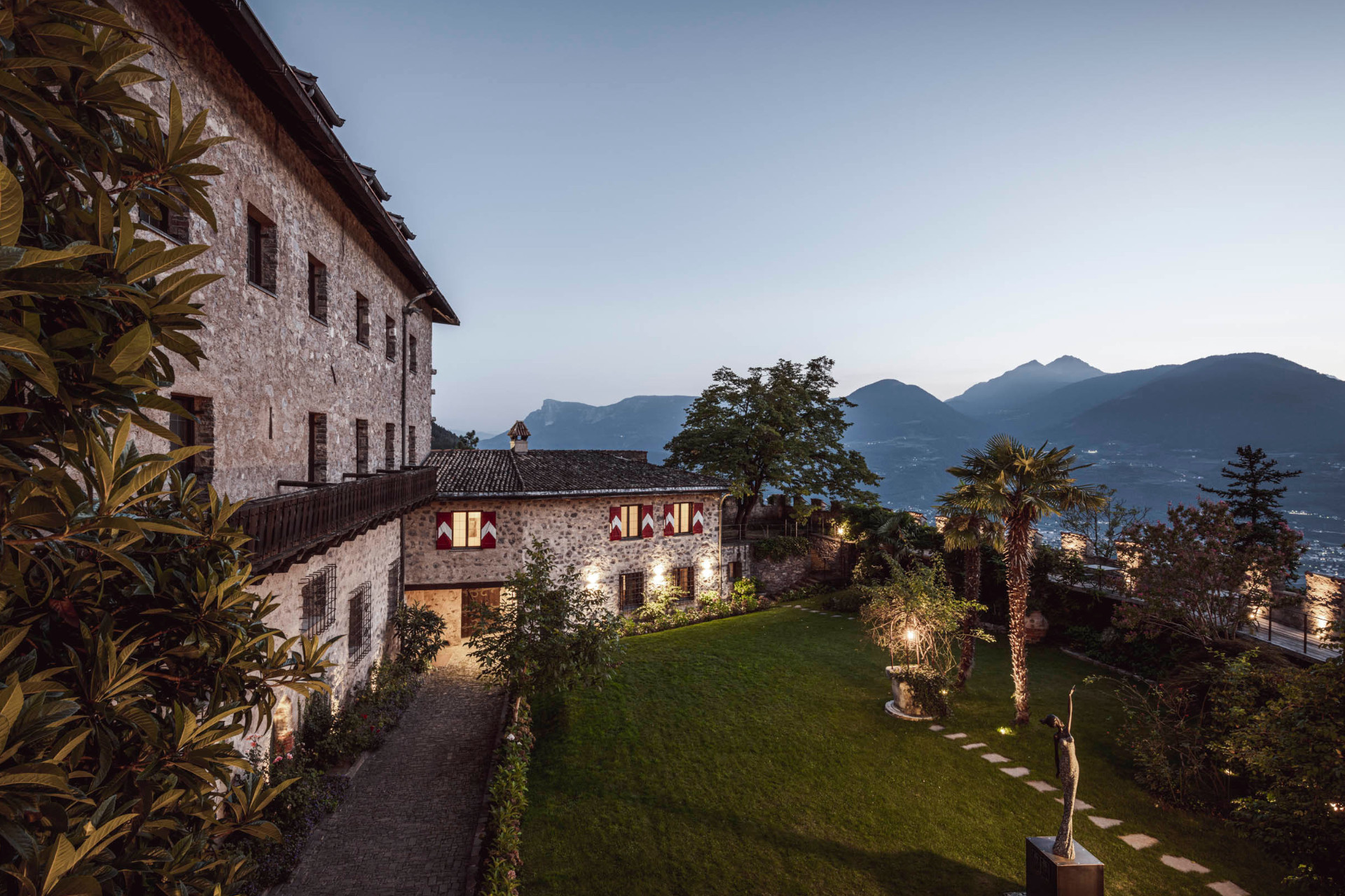Historic Castel Fragsburg at dusk, featuring a landscaped courtyard, mountains in the background, and illuminated greenery.