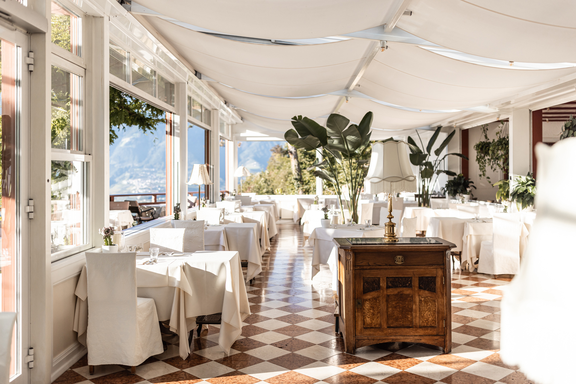 Elegant dining area in Castel Fragsburg's Orangerie, featuring white tablecloths, plants, and scenic views.