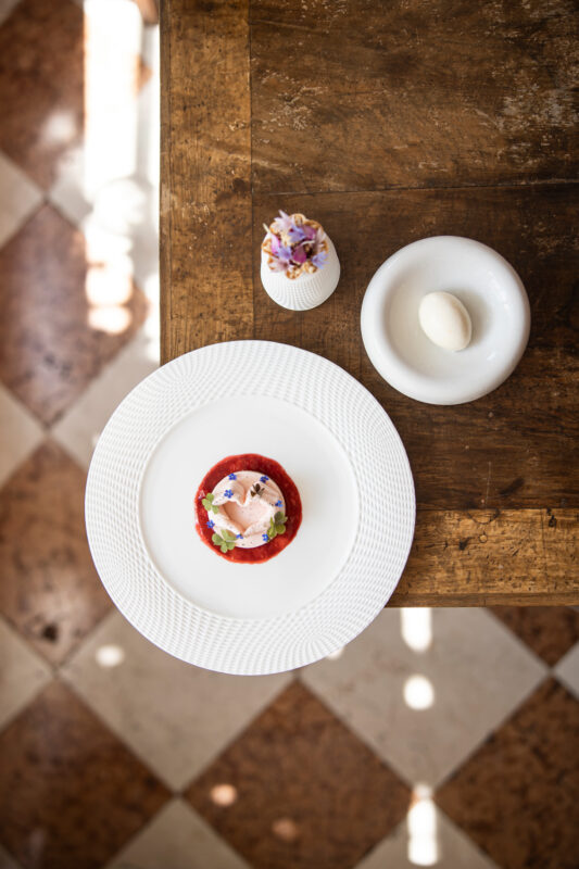 Dessert on a white plate with colorful toppings, accompanied by a small bowl and a flower arrangement, on a wooden table.