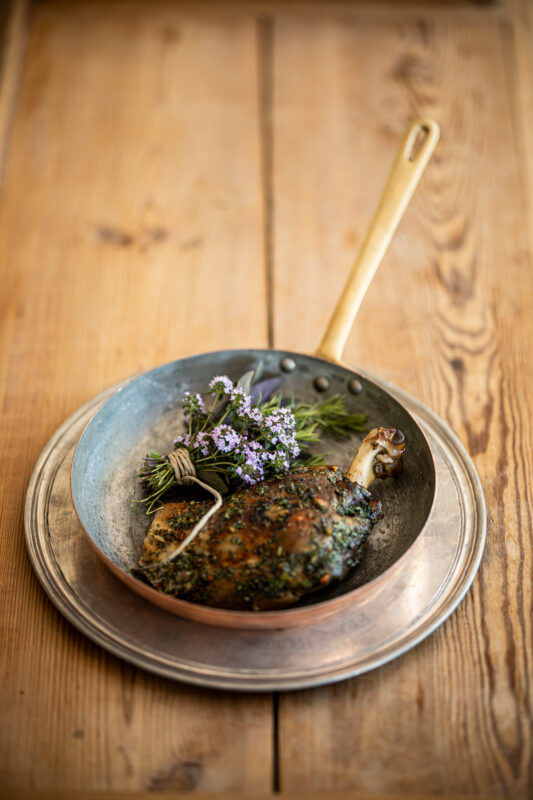 Roasted meat dish garnished with herbs, served in a copper pan on a wooden table.