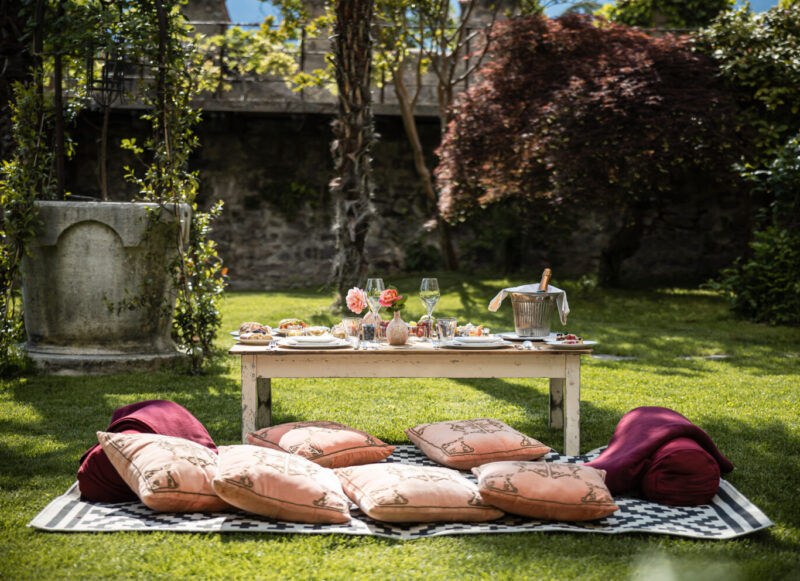 A cozy outdoor picnic setup with a low table, cushions, and a spread of food and drinks in a lush garden.