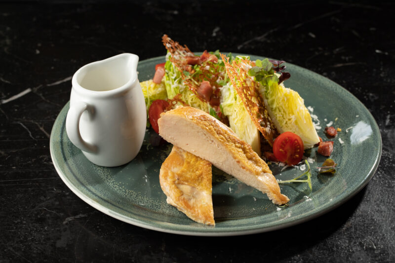 Ceasar salad with sliced chicken, cherry tomatoes, and a small jug of dressing on a green plate.