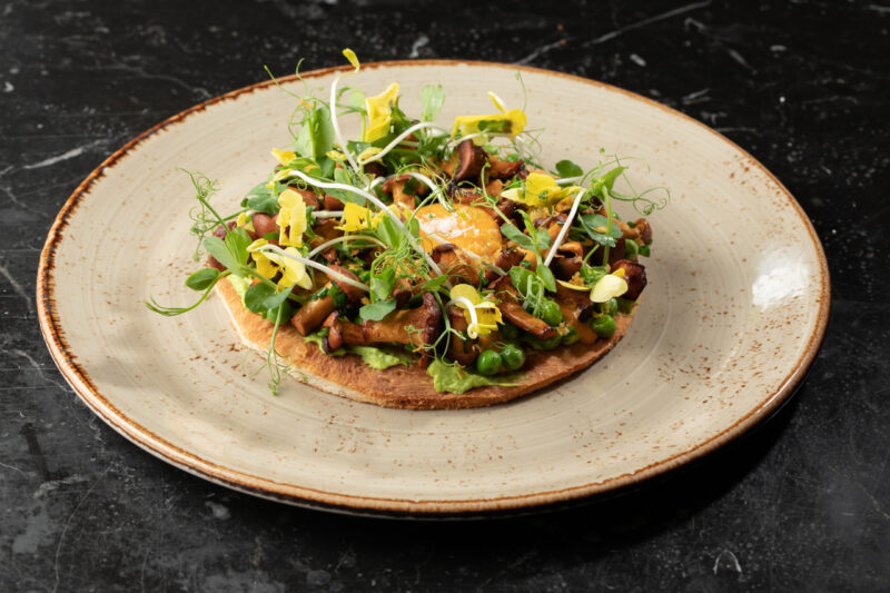 A plate of flatbread topped with sautéed chanterelle mushrooms, peas, and microgreens, garnished with yellow flowers.
